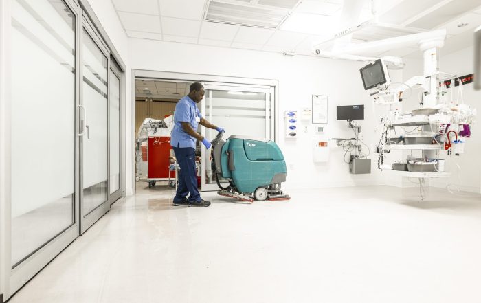 A caregiver operates a large, motorized floor cleaner in a hospital room.