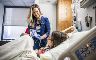 A Cleveland Clinic nurse in blue scrubs and gloves holds a medical device while caring for a patient lying in a hospital bed.