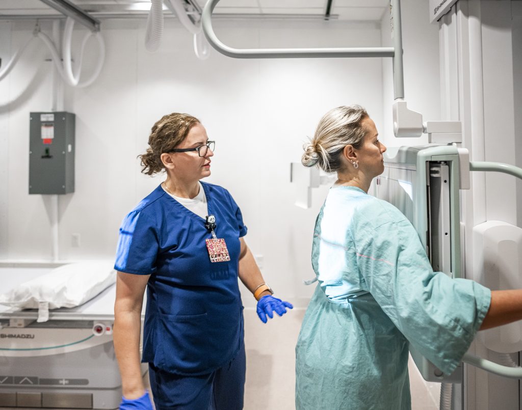 A Cleveland Clinic nurse in blue scrubs assists a patient in a hospital gown positioning for an imaging scan.