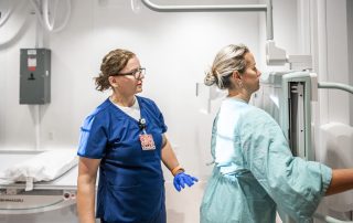 A Cleveland Clinic nurse in blue scrubs assists a patient in a hospital gown positioning for an imaging scan.