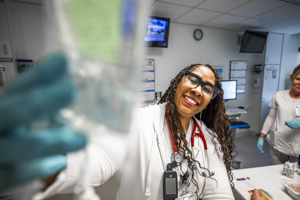 A smiling Cleveland Clinic nurse wearing glasses and a stethoscope adjusts an IV bag in a hospital unit.