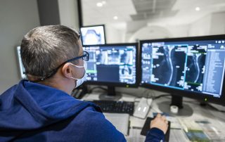 A Cleveland Clinic radiology specialist wearing a mask reviews patient scans on dual computer monitors in a control room.