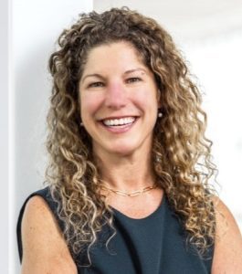 Smiling woman with curly hair wearing a sleeveless dark dress in a bright indoor setting.