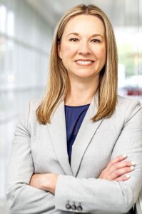 Professional woman smiling with arms crossed in a bright office hallway.