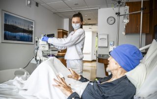 A Cleveland Clinic nurse in a mask and gloves adjusts medical equipment while an older patient wearing a blue hat rests in a hospital bed.