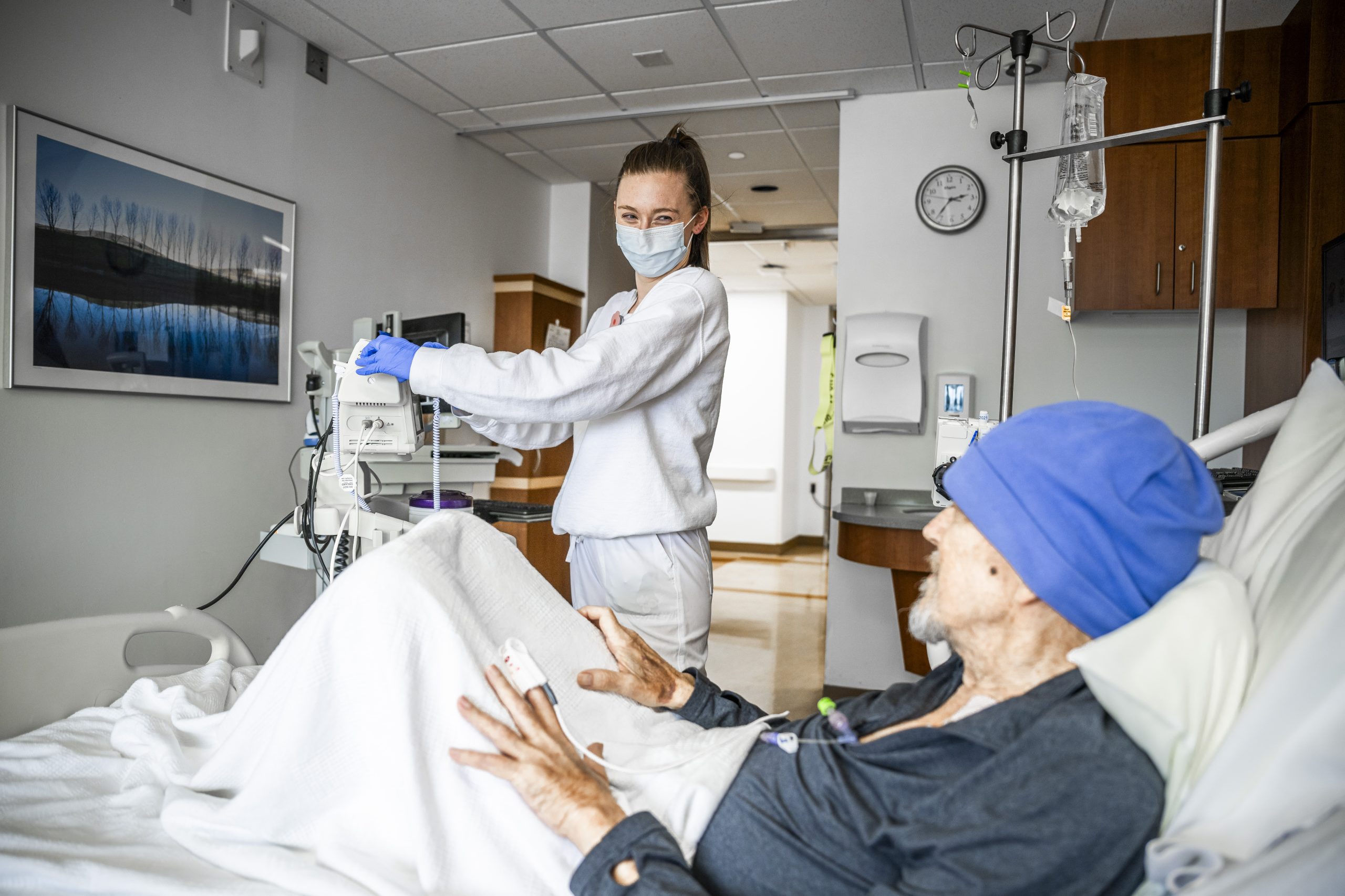 A Cleveland Clinic nurse in a mask and gloves adjusts medical equipment while an older patient wearing a blue hat rests in a hospital bed.