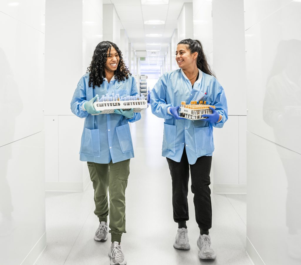 Two Cleveland Clinic laboratory staff in blue coats and gloves walk down a hallway smiling, each carrying trays of test tubes.