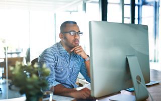 A man wearing glasses and a checkered shirt works at a computer in a modern office setting.