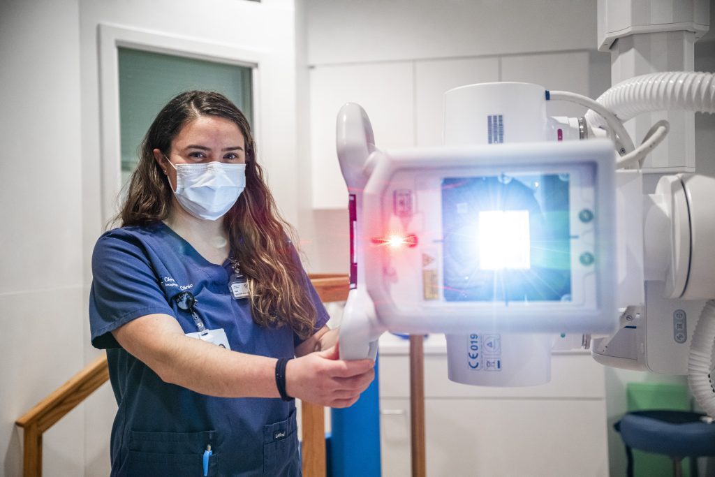 A Cleveland Clinic radiology technologist wearing scrubs and a mask positions an X-ray machine, with the device’s light shining toward the patient area.