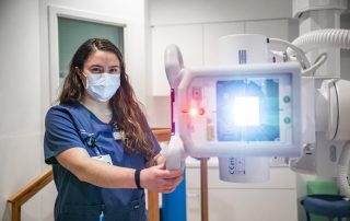 A Cleveland Clinic radiology technologist wearing scrubs and a mask positions an X-ray machine, with the device’s light shining toward the patient area.