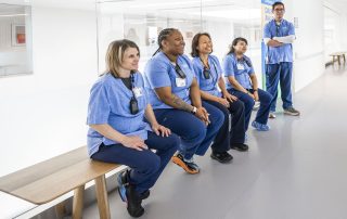 A group of Cleveland Clinic employees in blue scrubs sit together on a bench in a hospital hallway, smiling and talking.