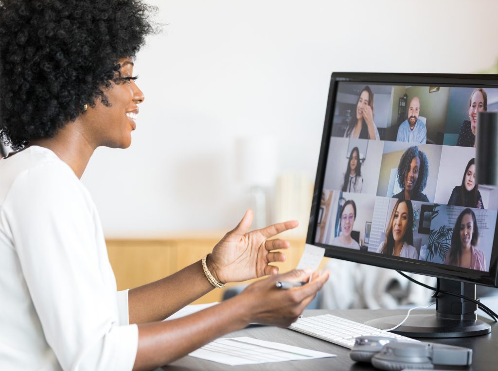 A woman smiles and gestures while leading a virtual meeting, with multiple colleagues visible on her computer screen in a video conference grid.