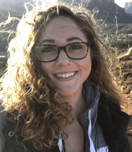 Smiling woman with curly hair and glasses outdoors in sunny weather.