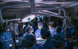 A male healthcare worker in blue scrubs and a face mask assists an older male patient with a breathing test using respiratory equipment in a clinical setting.