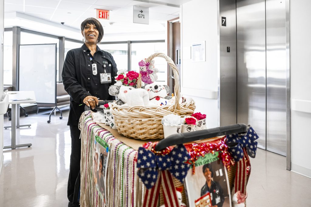 A smiling Cleveland Clinic employee pushes a decorated cart with baskets of flowers and stuffed animals in a hospital hallway.