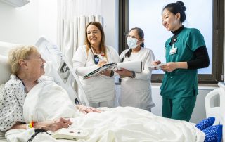A group of Cleveland Clinic nurses and staff stand beside a hospital bed, smiling and talking with an older patient.