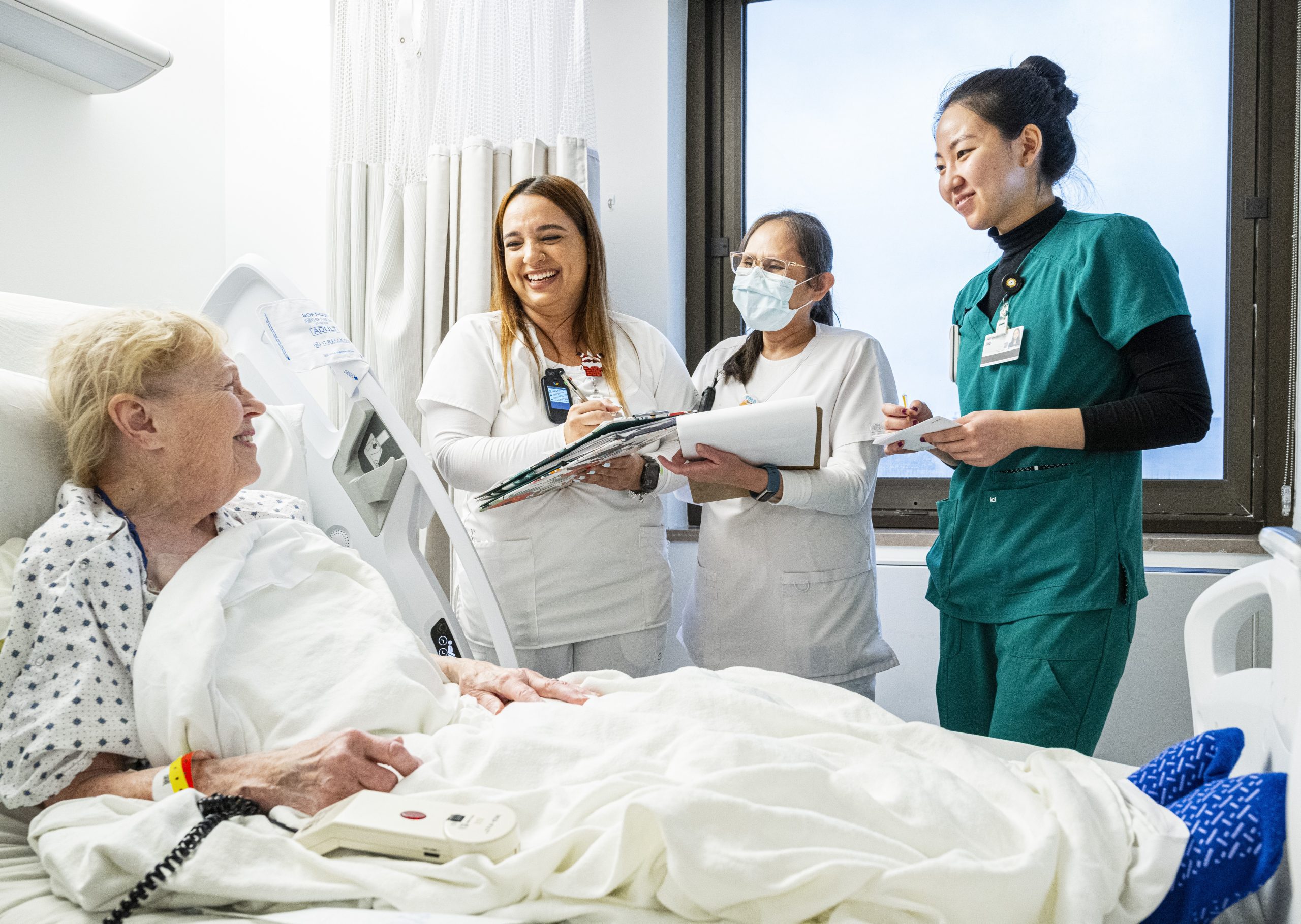 A group of Cleveland Clinic nurses and staff stand beside a hospital bed, smiling and talking with an older patient.