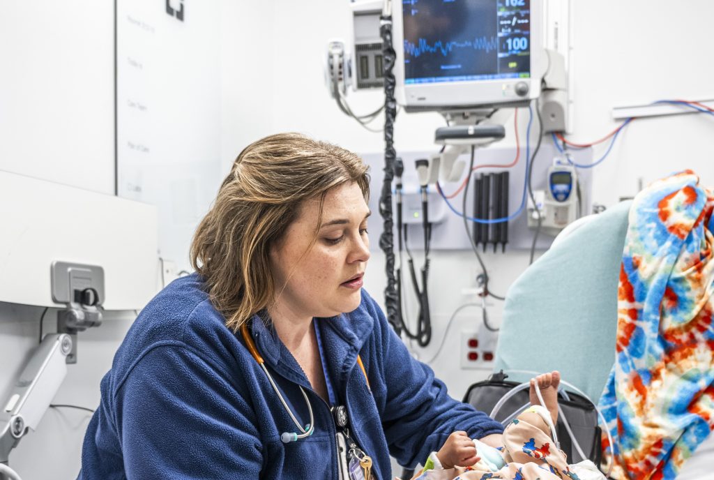 A woman nurse wearing a stethoscope holds an infant patient in a clinical setting with medical monitoring equipment behind them.