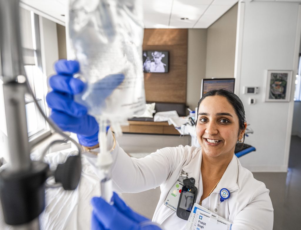 A smiling Cleveland Clinic nurse wearing gloves adjusts an IV fluid bag in a hospital room.
