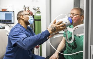 A male healthcare worker in blue scrubs and a face mask assists an older male patient with a breathing test using respiratory equipment in a clinical setting.