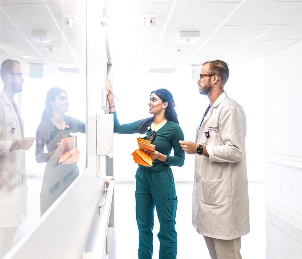 A Cleveland Clinic nurse in green scrubs writes on a whiteboard while collaborating with a doctor in a white coat in a hospital hallway.