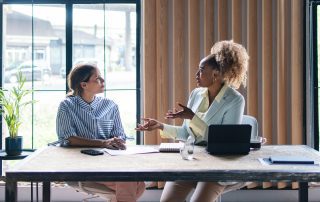 Two professional women sit across from each other at a desk in an office, engaged in a focused conversation with a laptop and documents in front of them.