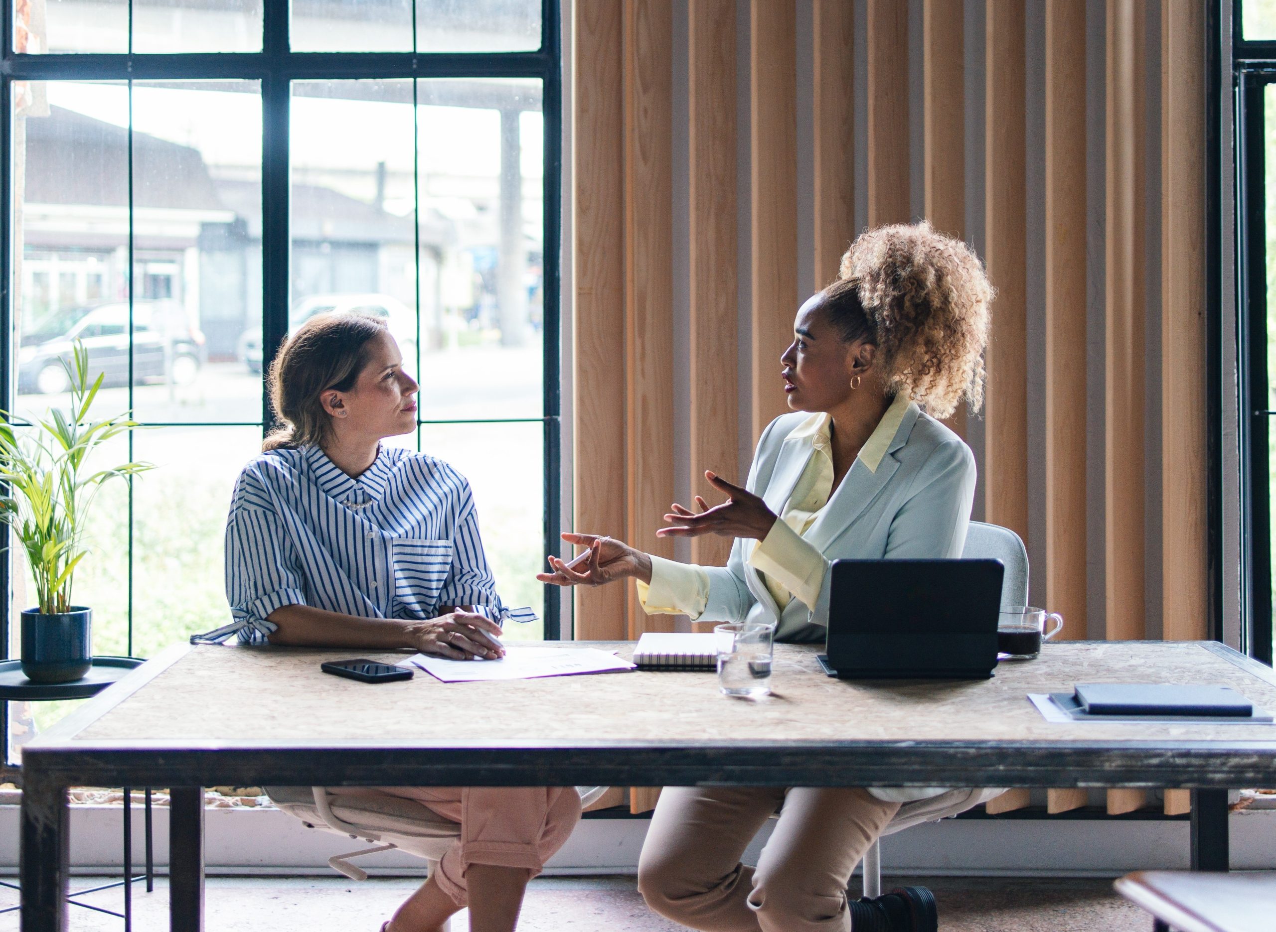 Two professional women sit across from each other at a desk in an office, engaged in a focused conversation with a laptop and documents in front of them.