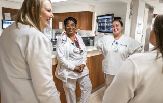 A group of Cleveland Clinic nurses in white scrubs stand together at a nurses’ station, smiling and talking with each other.
