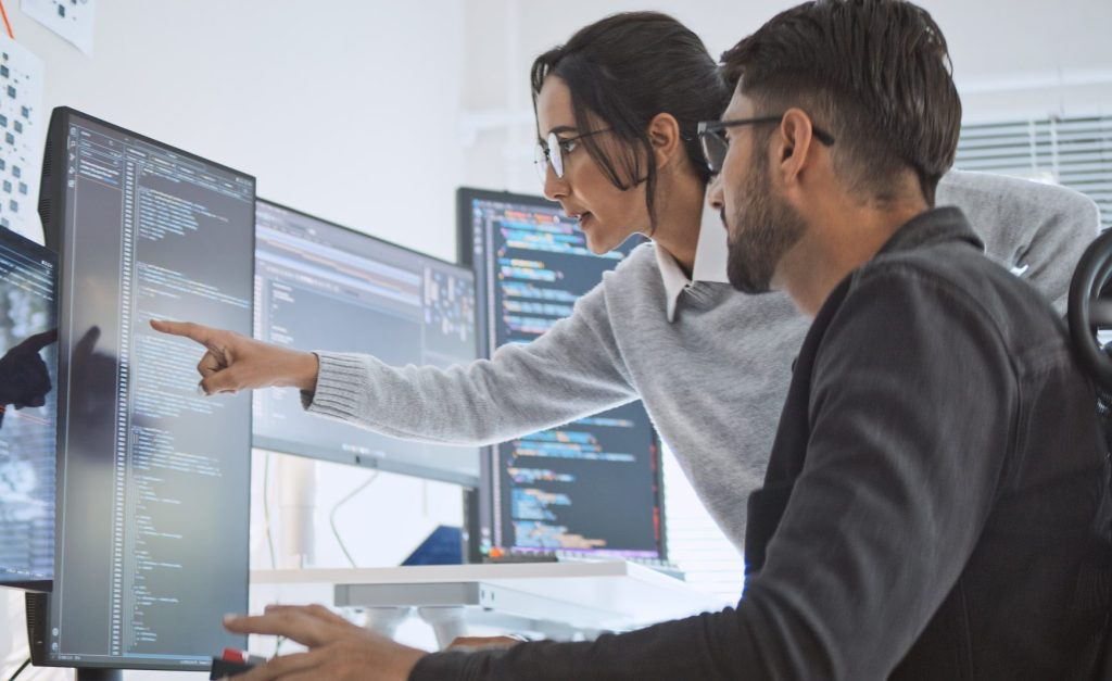 A woman points at code on a computer monitor while collaborating with a male colleague in an office setting with multiple screens displaying programming.