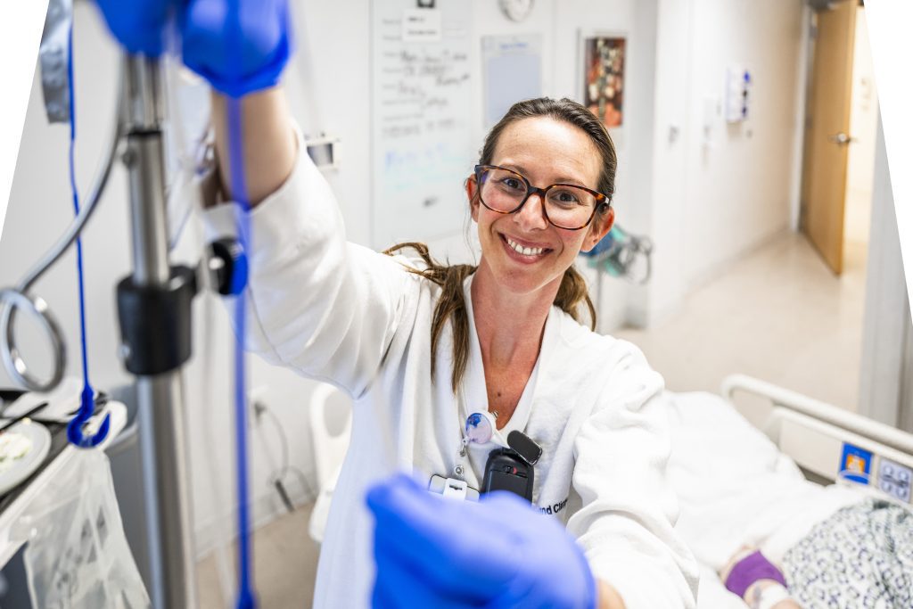 A smiling Cleveland Clinic nurse wearing glasses and gloves adjusts IV tubing beside a patient’s hospital bed.