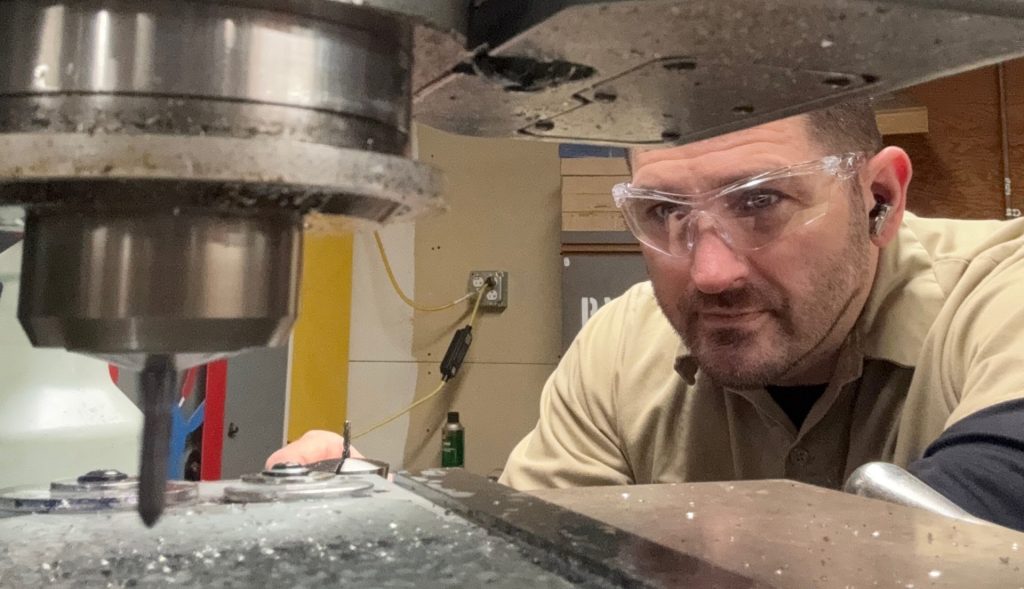 A man wearing safety glasses operates a large industrial milling machine, closely monitoring the drill bit cutting into metal.