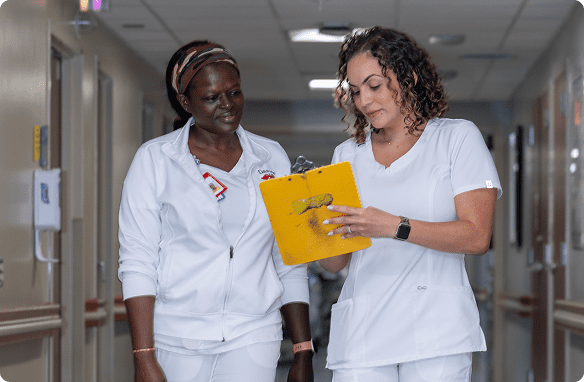 Two Cleveland Clinic nurses in white scrubs walk together in a hospital hallway while reviewing notes on a yellow clipboard.