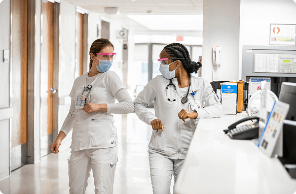Two Cleveland Clinic nurses in white scrubs, masks, and face shields greet each other with an elbow bump in a hospital hallway.