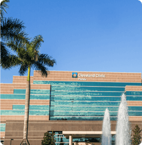 Exterior view of the Cleveland Clinic Florida building with palm trees and a fountain in front under a clear blue sky.