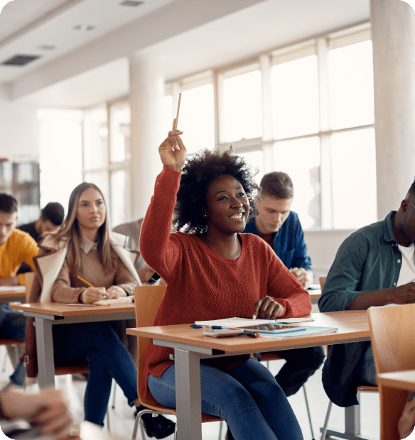 A student in a red sweater raises her hand while sitting at a desk in a classroom, surrounded by other students taking notes.