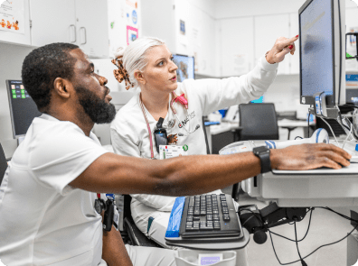 Two Cleveland Clinic nurses in white scrubs sit at a workstation, reviewing information on a computer as one points to the screen.