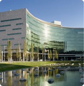 The glass-fronted Cleveland Clinic main building in Ohio reflects the sky and trees, with a landscaped pond and stones in the foreground.