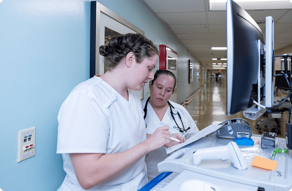 Two Cleveland Clinic nurses in white scrubs review notes together at a workstation in a hospital hallway.