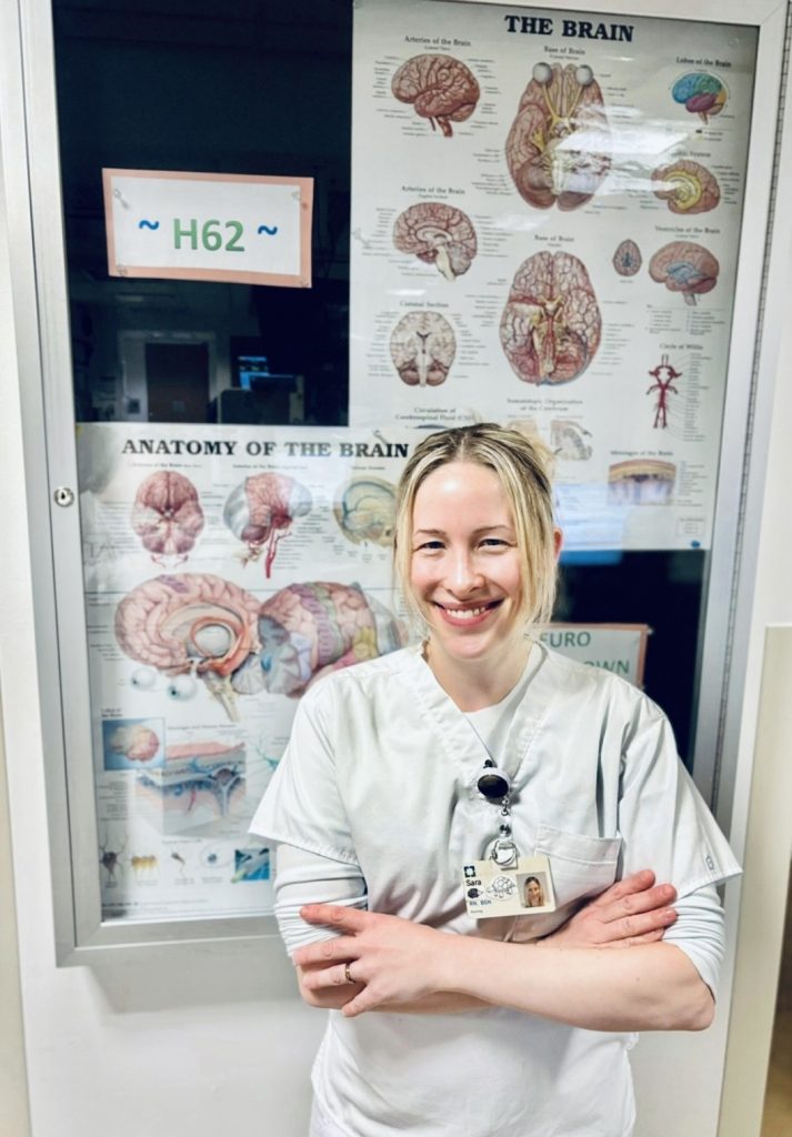 Sara wearing a Cleveland Clinic scrub top stands in front of a wall poster detailing brain anatomy.